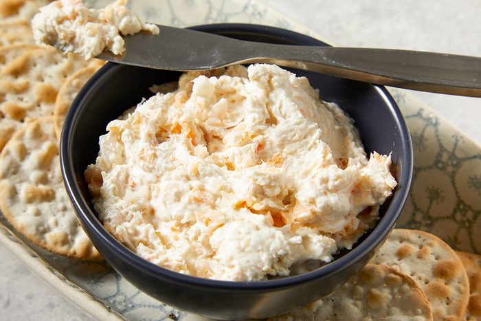 A bowl of creamy cheese spread with a knife resting on top, surrounded by round crackers on a patterned tray.