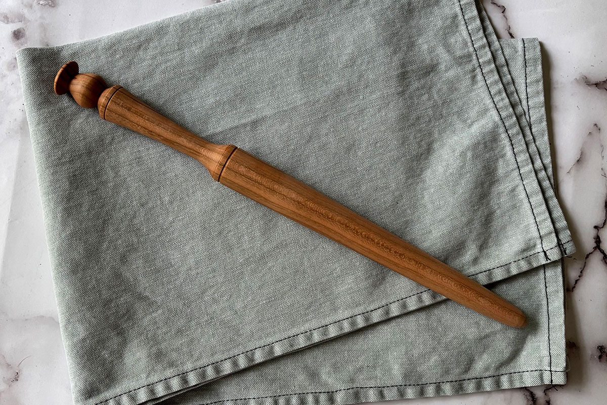 A wooden French rolling pin with tapered ends rests diagonally on a folded, light gray cloth atop a marble surface.