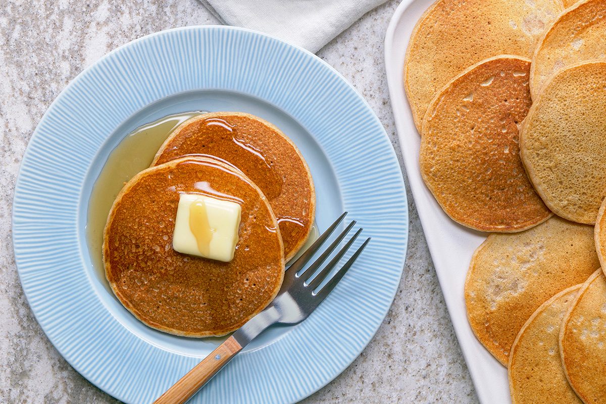 Two pancakes with a pat of butter and syrup on a blue plate with a fork, next to a tray of more pancakes on a light surface.