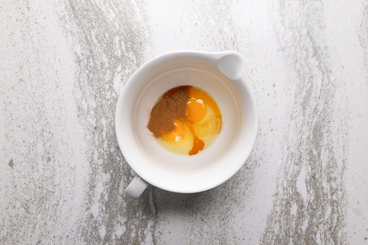 A white mixing bowl on a light marble surface contains an egg yolk, egg white, and brown spices ready to be mixed.