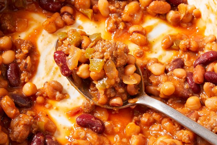 A close-up of a spoonful of chili with ground meat, red kidney beans, white beans, green peppers, and tomato sauce, surrounded by more chili in a dish.