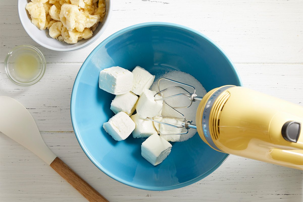 A yellow hand mixer in a blue bowl with cream cheese cubes and sugar, with a spatula, a small bowl of lemon juice, and a bowl of butter pieces nearby on a white surface.