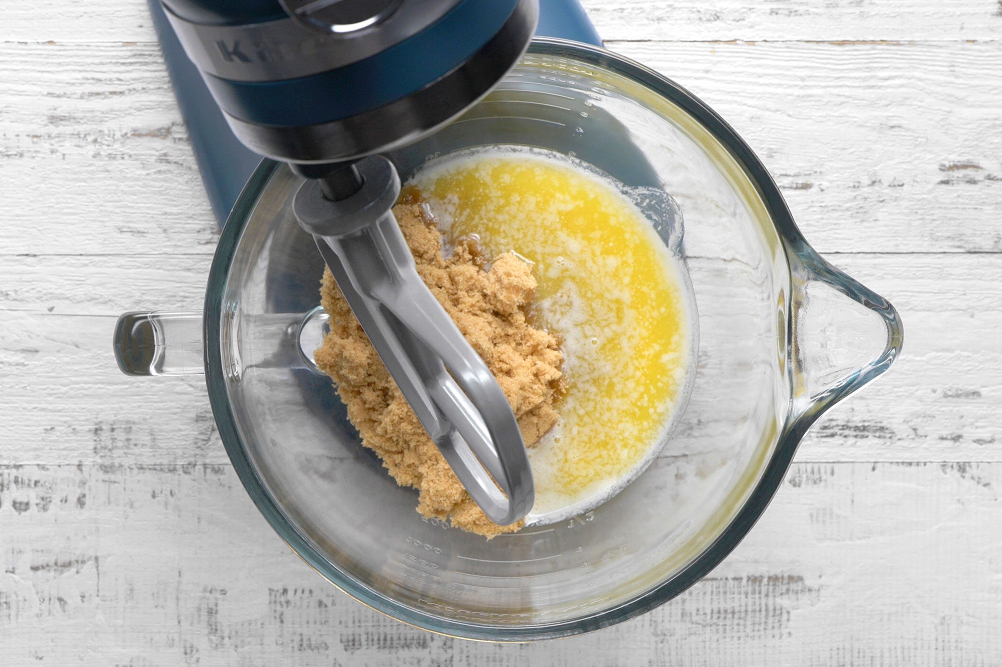 Overhead view of a stand mixer with a glass bowl containing melted butter and brown sugar, ready to be mixed, on a white wooden surface