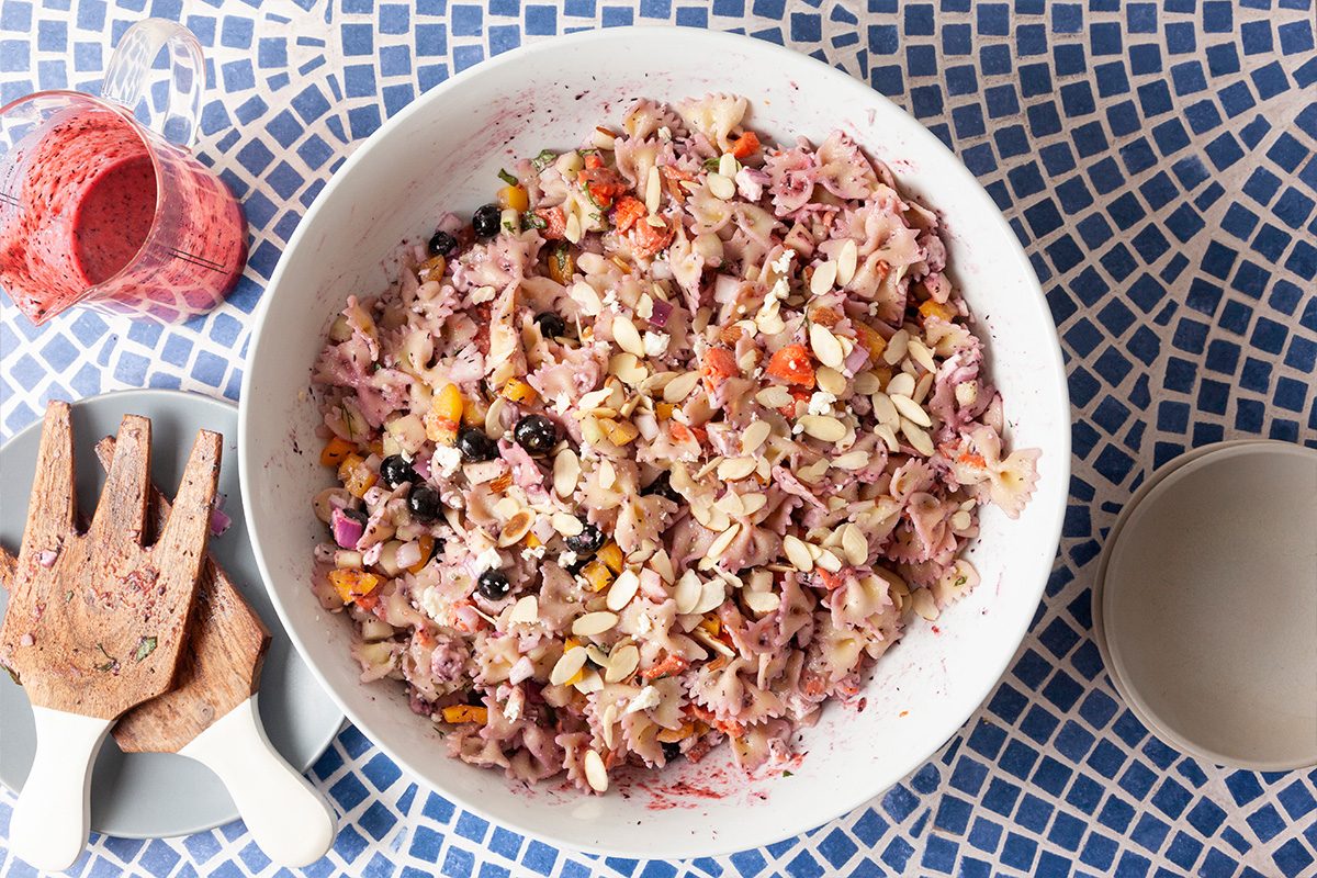 A bowl of mixed salad with rice, nuts, dried fruits, and feta cheese sits on a blue and white mosaic table, with wooden salad servers and a small glass of pink dressing nearby.