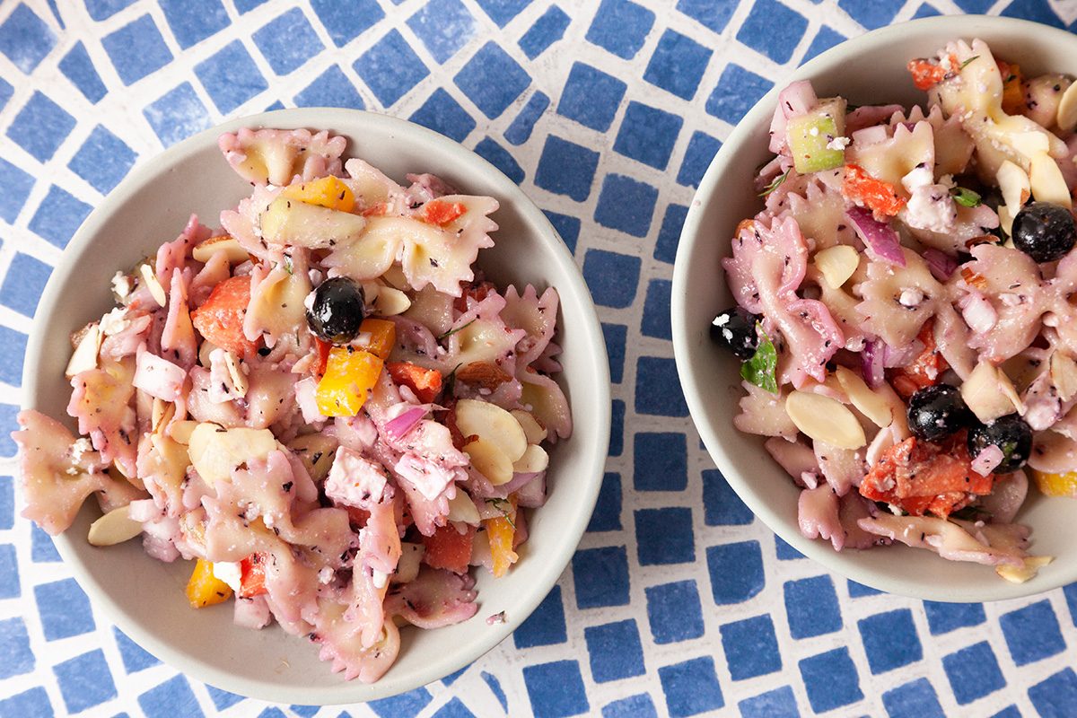 Two bowls of colorful pasta salad with bowtie pasta, black olives, feta cheese, and chopped vegetables, placed on a blue and white mosaic-patterned surface.