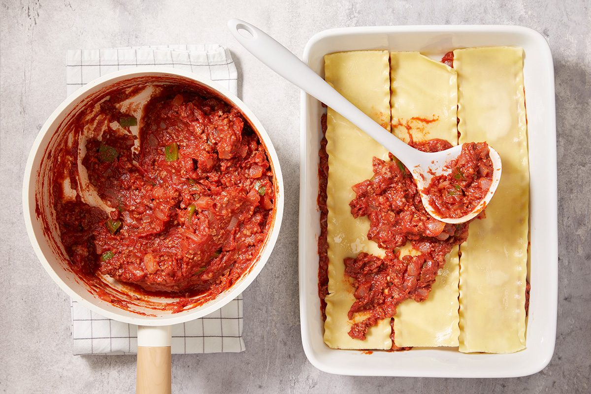 A saucepan of chunky red tomato meat sauce sits beside a baking dish with uncooked lasagna noodles. A white spoon spreads sauce over the noodles, preparing a lasagna on a light countertop.