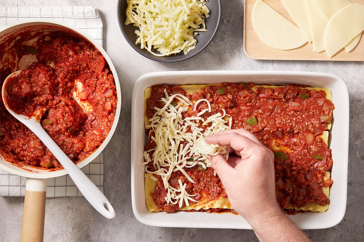 A hand sprinkles shredded cheese over a layer of lasagna with tomato sauce in a white baking dish. Nearby are a pot of sauce, a bowl of shredded cheese, and slices of cheese on a cutting board.
