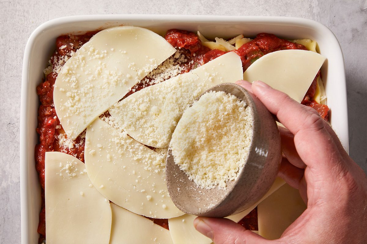 A hand sprinkles grated Parmesan cheese from a small bowl onto a layered lasagna topped with tomato sauce and slices of mozzarella cheese in a white baking dish.