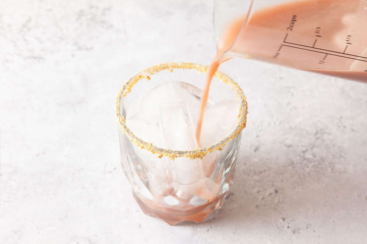 A pink drink is being poured from a measuring cup into a glass with large ice cubes and a rim coated in gold-colored sugar or salt. The background is a light, textured surface.