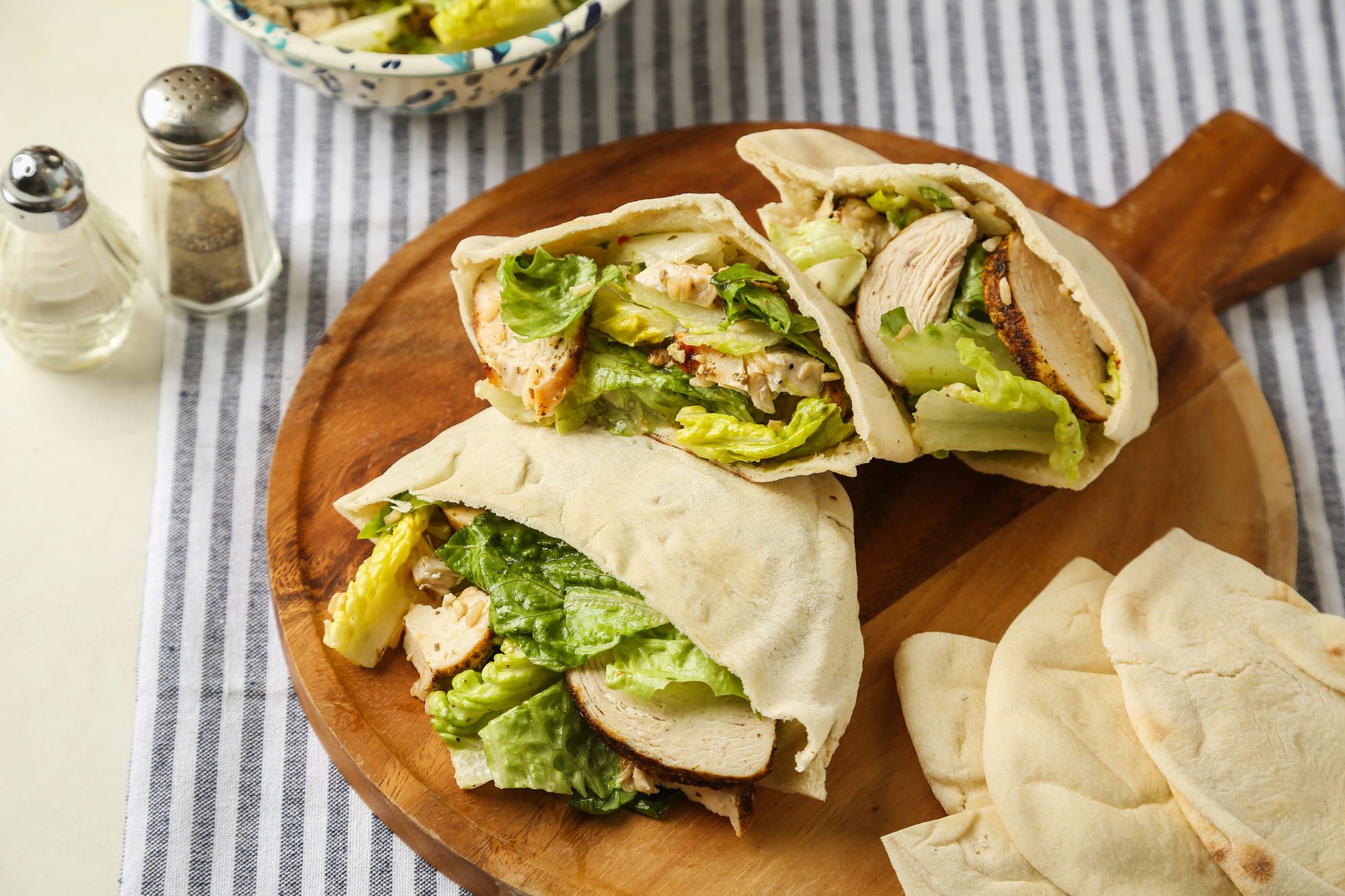 Close view of Chicken Caesar Pitas; served on a round wooden tray; accompanied by salt and pepper shakers; all set on a grey and white linen cloth