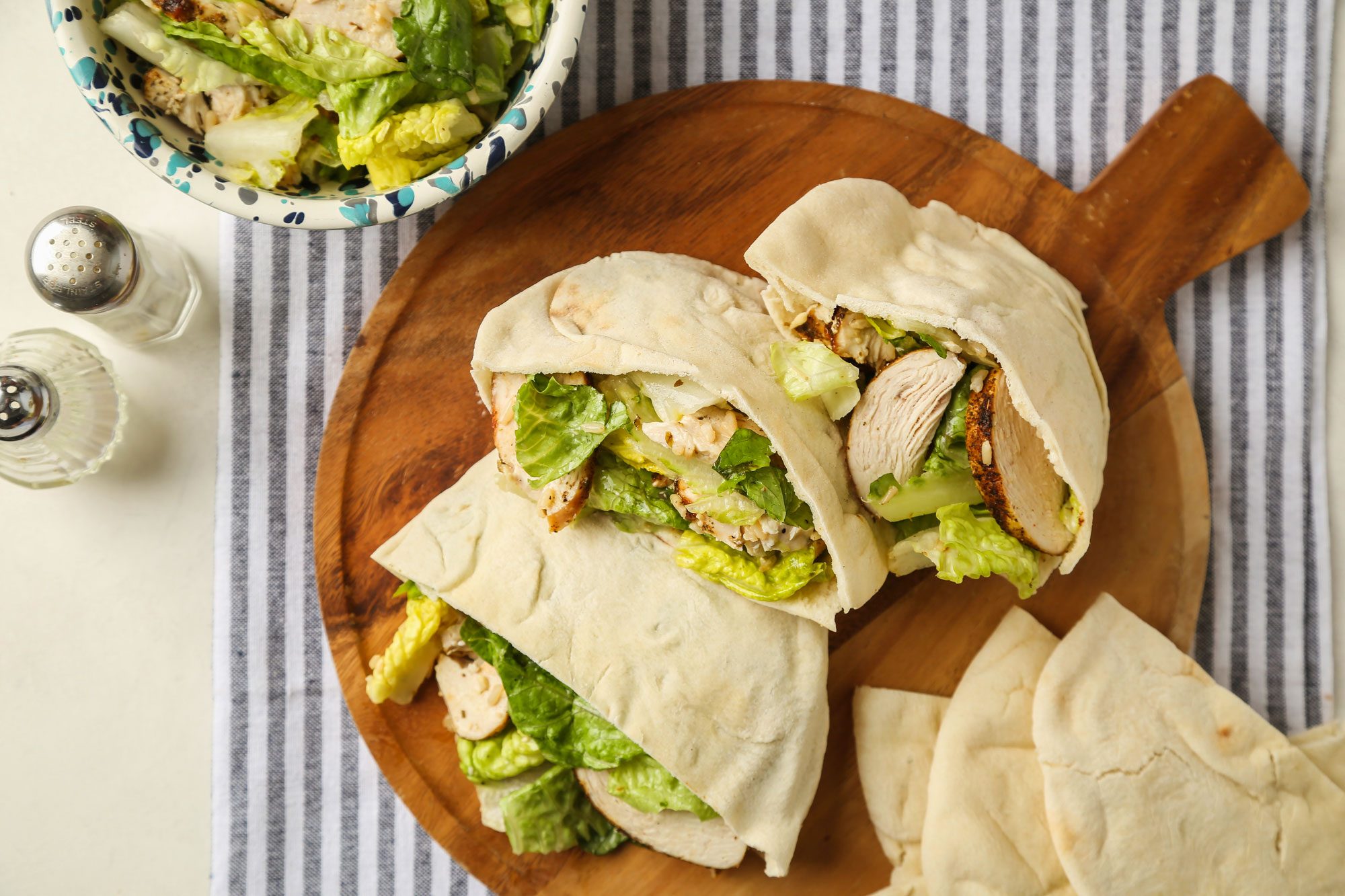 Overhead of Chicken Caesar Pitas; served on a round wooden tray; accompanied by salt and pepper shakers; all set on a grey and white linen cloth