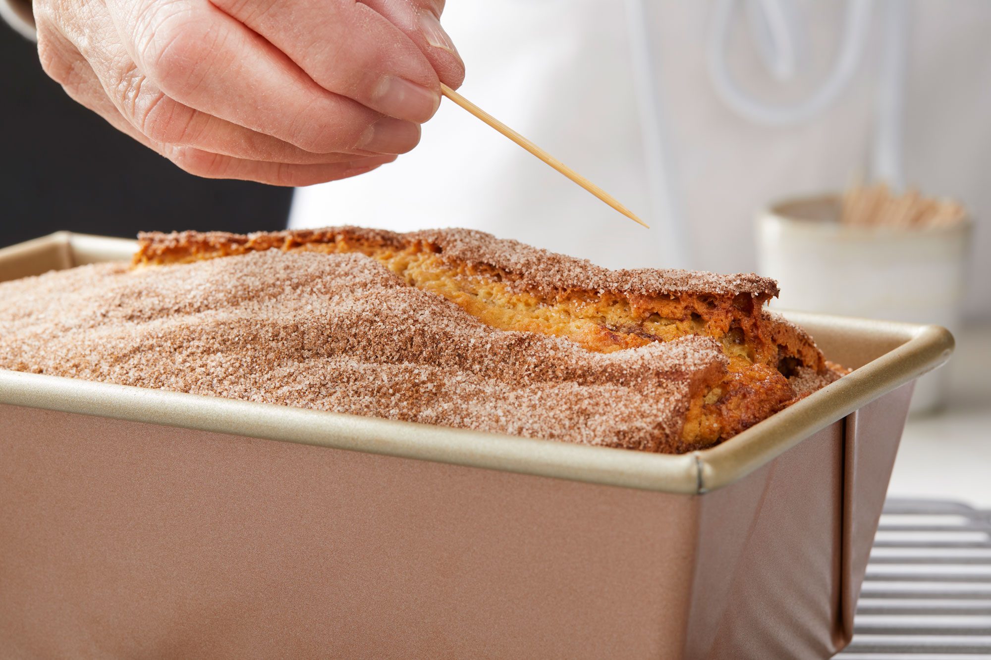 a closeup of a person testing the doneness of a freshly baked loaf in a metal pan by inserting a wooden skewer into the bread’s center, The loaf is topped with a layer of cinnamon sugar