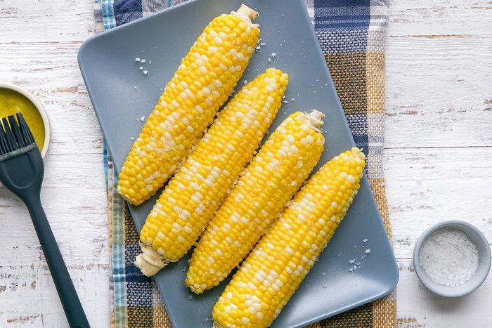 Four ears of corn on the cob are arranged on a gray plate, sprinkled with salt. A basting brush and a small dish of butter are on the side, all set on a plaid cloth and white wooden table.