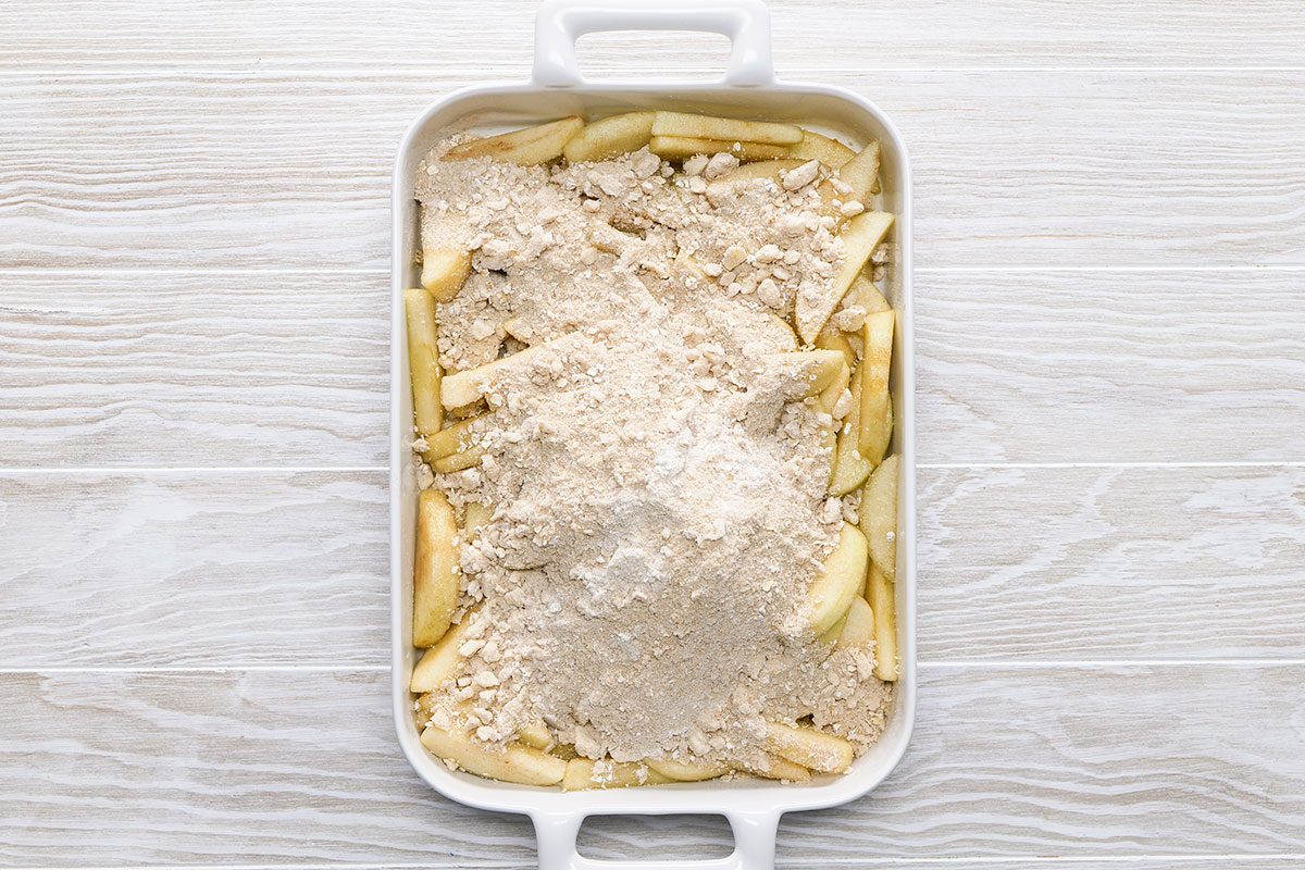 A white baking dish filled with sliced apples topped with an even layer of dry crumble mixture, ready to be baked, sits on a light wood surface.