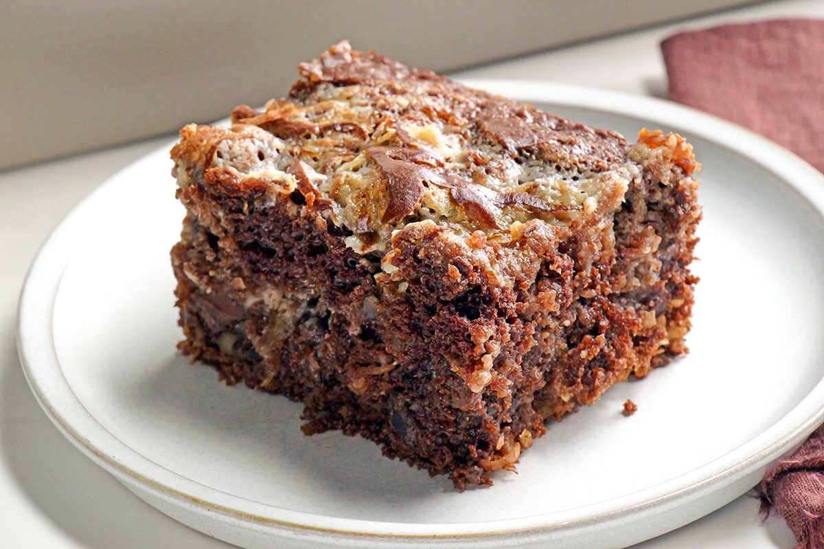 A square piece of chocolate and cream cheese marble cake sits on a white plate, showing a swirled pattern of dark chocolate and creamy layers. A brown napkin is partly visible beside the plate.