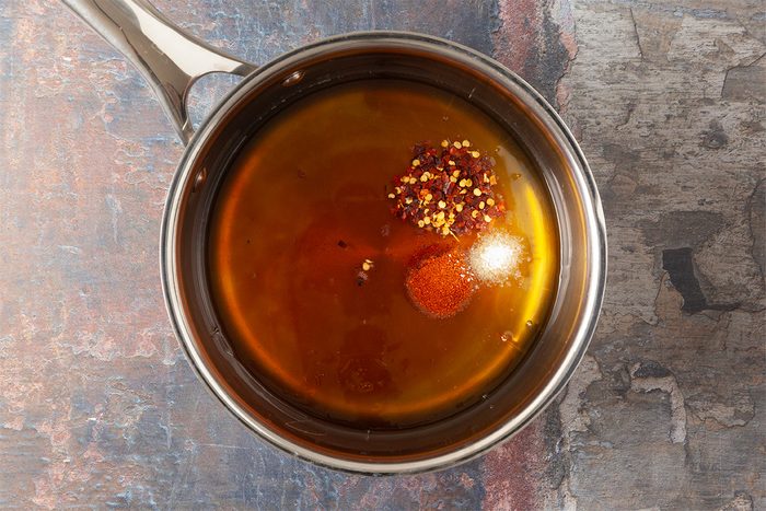 A metal saucepan containing a brown liquid with visible red pepper flakes and a small mound of white granules, viewed from above on a rustic surface.