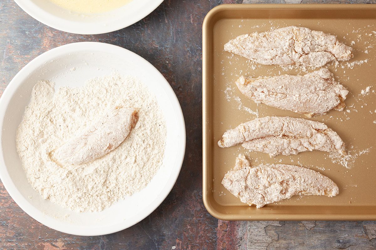 A plate of flour with a chicken strip being coated sits next to a baking sheet lined with several breaded chicken strips, ready to be cooked.