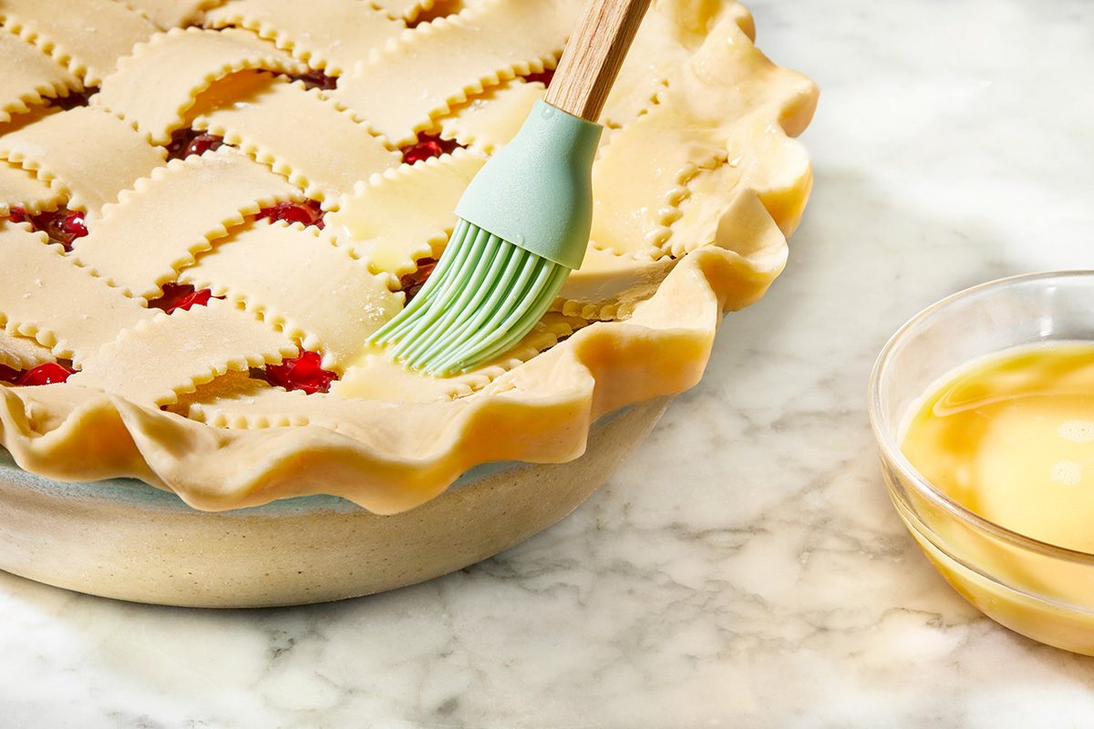 A pie with a decorative lattice crust is being brushed with an egg wash using a silicone pastry brush. A small bowl of egg wash sits to the side on a marble countertop.