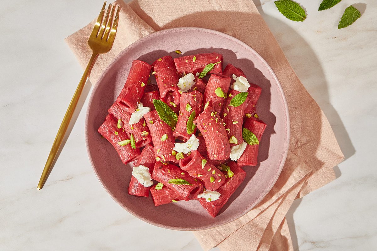 A pink bowl of rolled beet pasta topped with dollops of ricotta cheese, fresh mint leaves, and sprinkled nuts, placed on a pink napkin with a gold fork beside it on a marble surface.