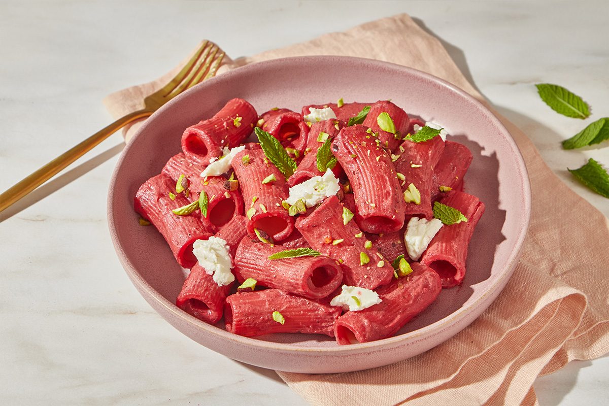 A pink bowl filled with bright pink rigatoni pasta, garnished with crumbled cheese, pistachios, and fresh mint leaves, sits on a matching pink napkin with a gold fork beside it.