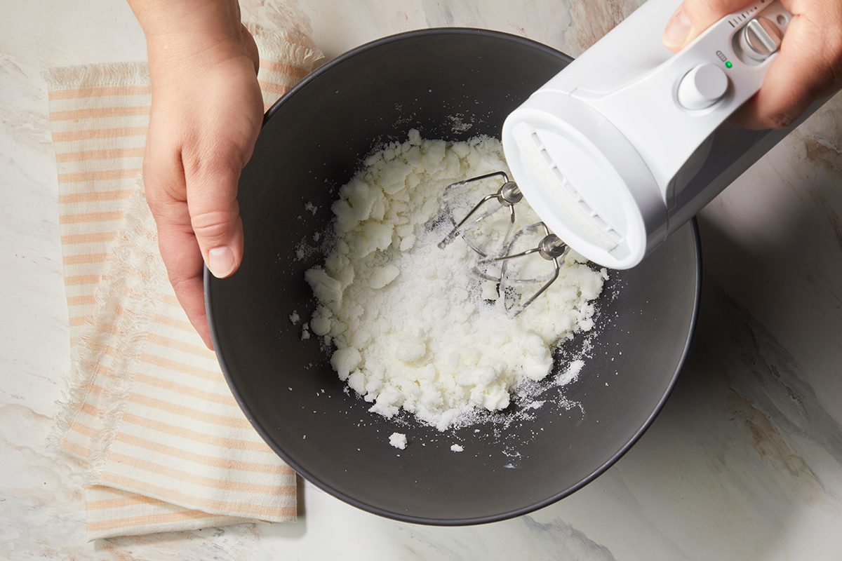 A person uses an electric hand mixer to blend white ingredients in a black bowl on a marble surface, with a folded striped cloth nearby.