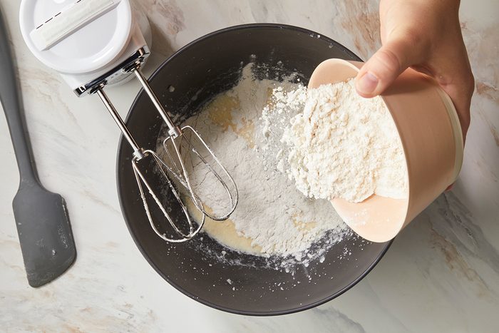 A hand pours flour from a bowl into a mixing bowl with a hand mixer and some flour already inside. A spatula and a container with a lid are on the countertop nearby.