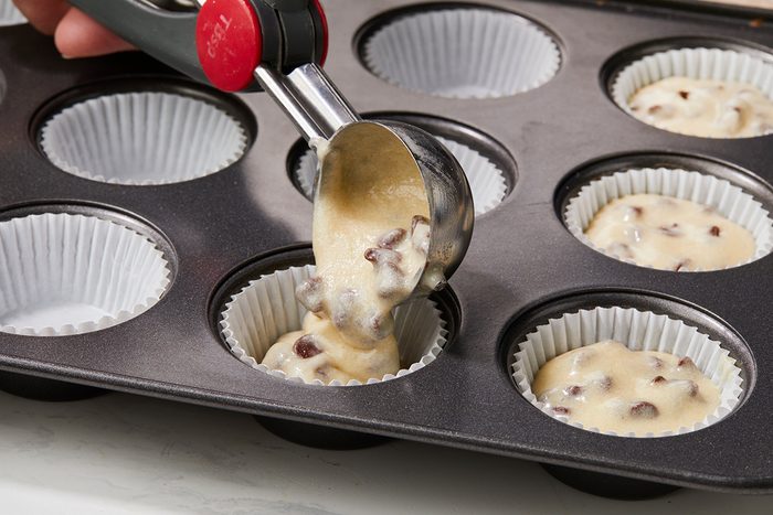 A hand uses a metal scoop to fill a paper-lined muffin tin with chocolate chip muffin batter. Several cups are already filled, and the muffin pan sits on a light surface.