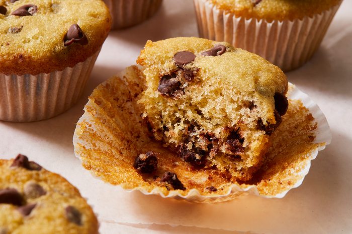 A close-up of chocolate chip muffins, with one muffin partially unwrapped and bitten to reveal its soft, moist texture and chocolate chips inside. Other whole muffins are visible in the background.