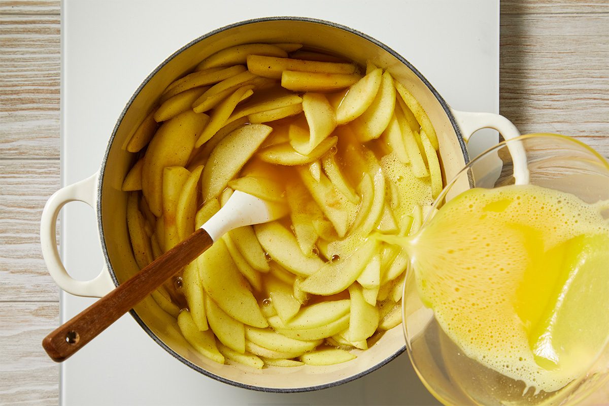 Sliced apples in a white pot are being mixed with a liquid mixture poured from a glass bowl, with a white spatula resting in the pot. The scene is set on a light wooden surface.