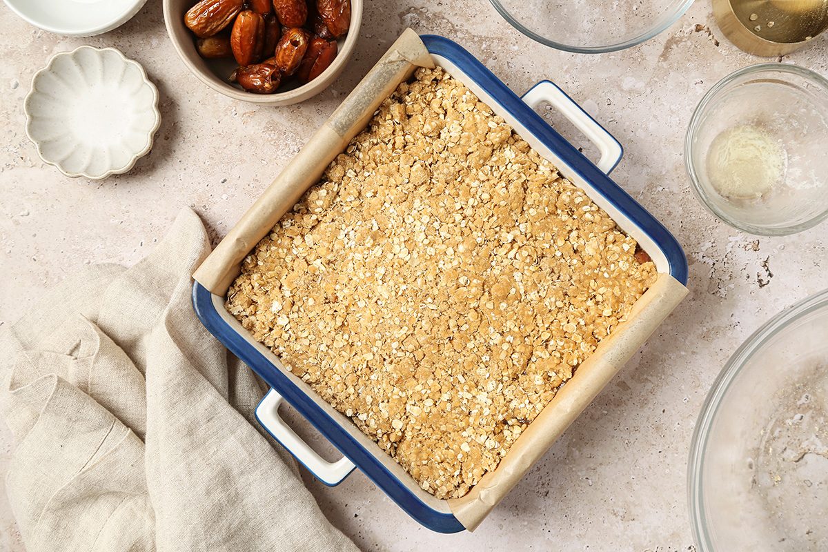 A square baking dish lined with parchment paper and filled with an unbaked crumbly oat mixture sits on a counter, surrounded by bowls with dates and baking ingredients.