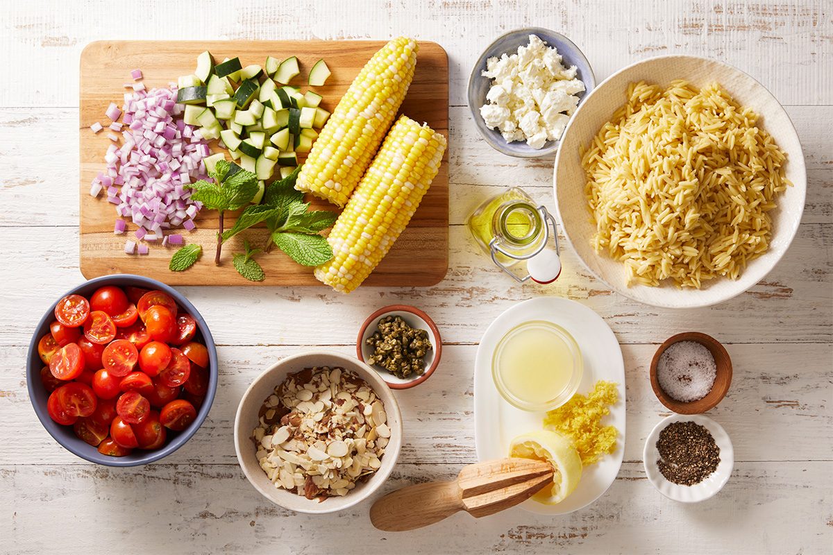 A top-down view of ingredients for a pasta dish, including chopped tomatoes, corn on the cob, diced zucchini, chopped onions, cooked orzo, feta cheese, herbs, olive oil, lemon juice, and seasonings on a white table.