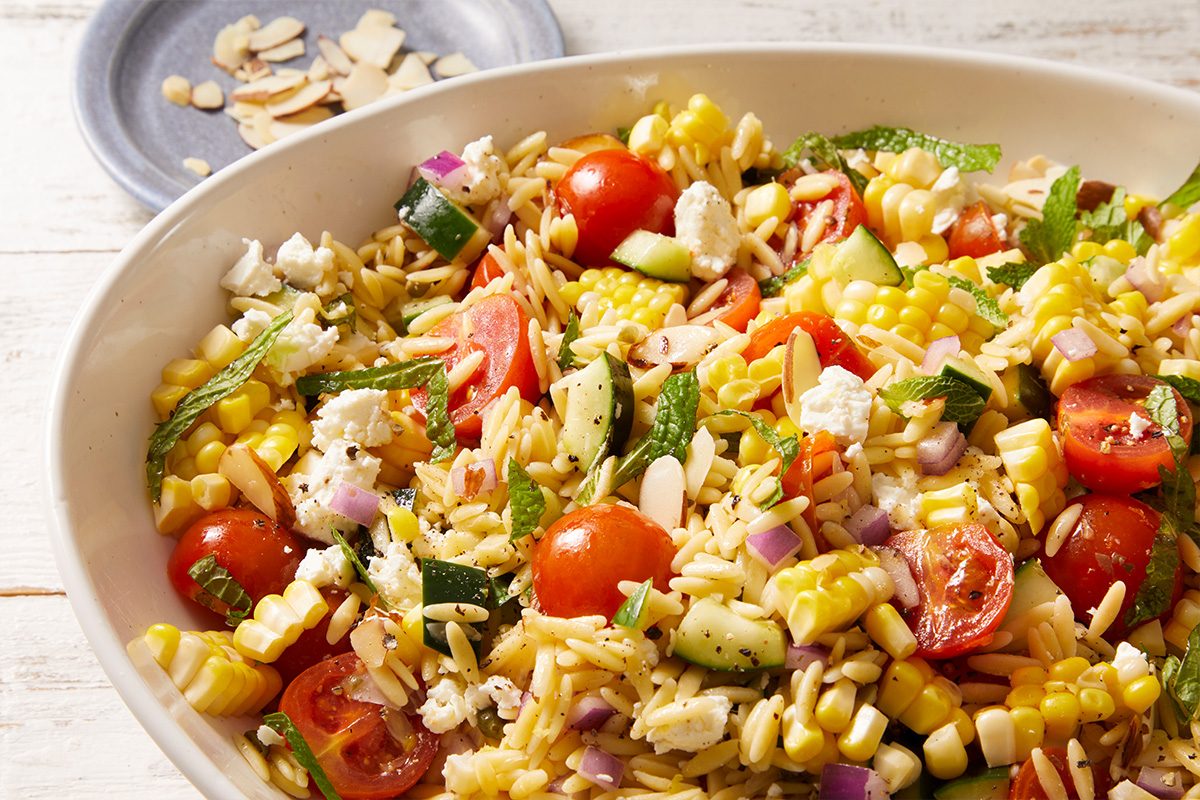 A bowl of orzo pasta salad with cherry tomatoes, corn, cucumber, red onion, feta cheese, sliced almonds, and fresh herbs, set on a light table with a small dish of almond slices nearby.