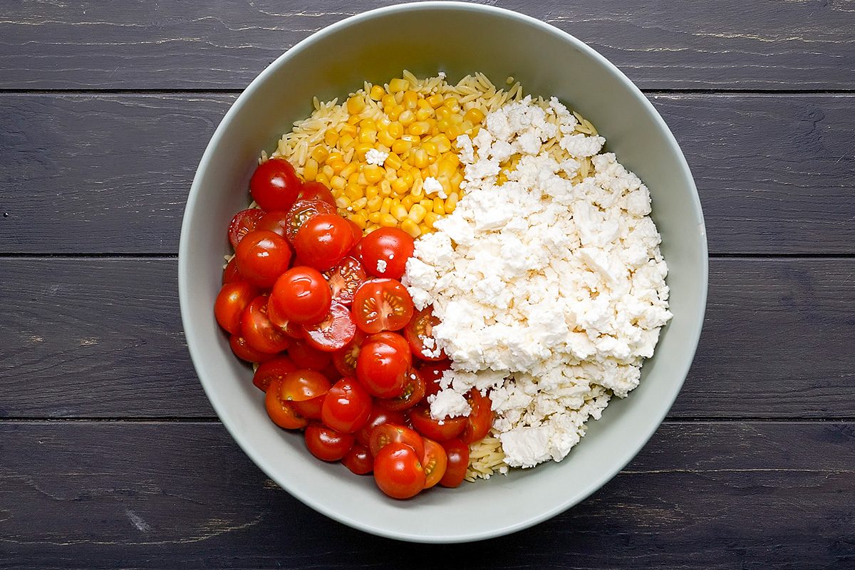 A bowl containing cooked orzo pasta, yellow corn, halved cherry tomatoes, and crumbled feta cheese, arranged in separate sections on a dark wooden surface.