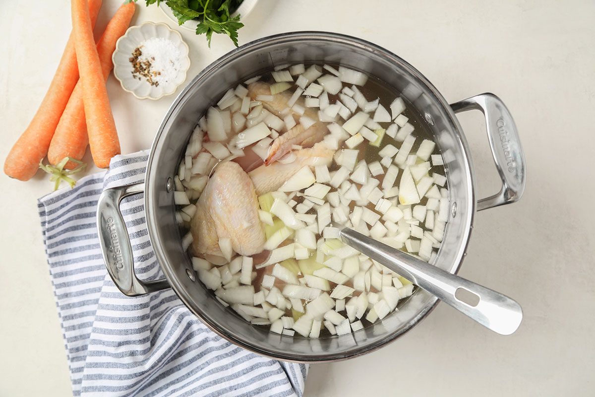 A pot filled with chopped onions and raw chicken pieces sits on a striped towel, with a spoon inside. Carrots, a bowl of salt and pepper, and fresh herbs are on the side on a light surface.