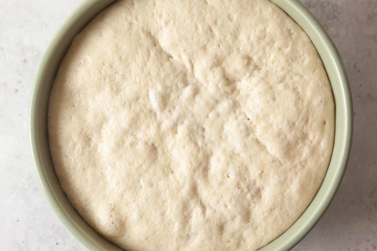 A bowl filled with risen bread dough, showing a smooth, slightly bumpy surface, indicating that it has been proofed and is ready for the next baking step.