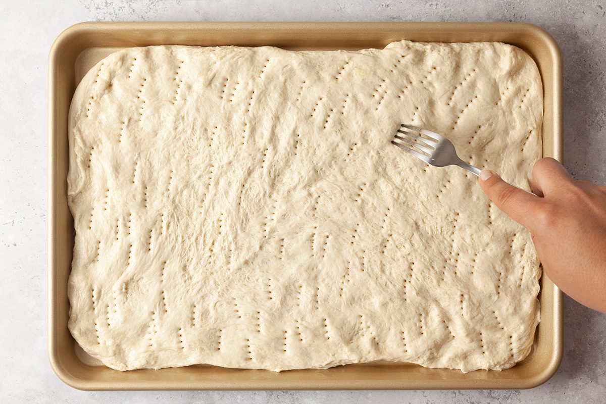 A hand uses a fork to poke holes in rectangular pizza dough spread evenly on a baking sheet.