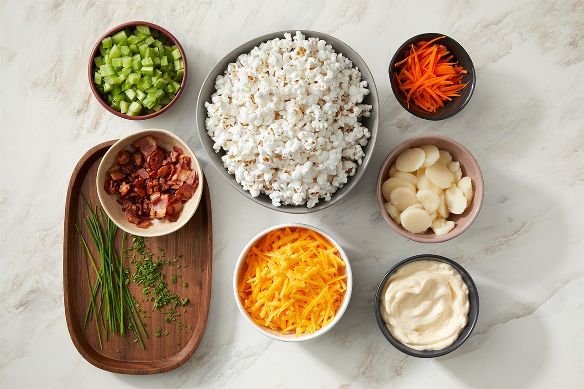 A variety of ingredients in bowls on a white surface, including popcorn, sliced celery, shredded carrots, bacon bits, water chestnuts, shredded cheddar cheese, chopped chives, and a bowl of mayonnaise.