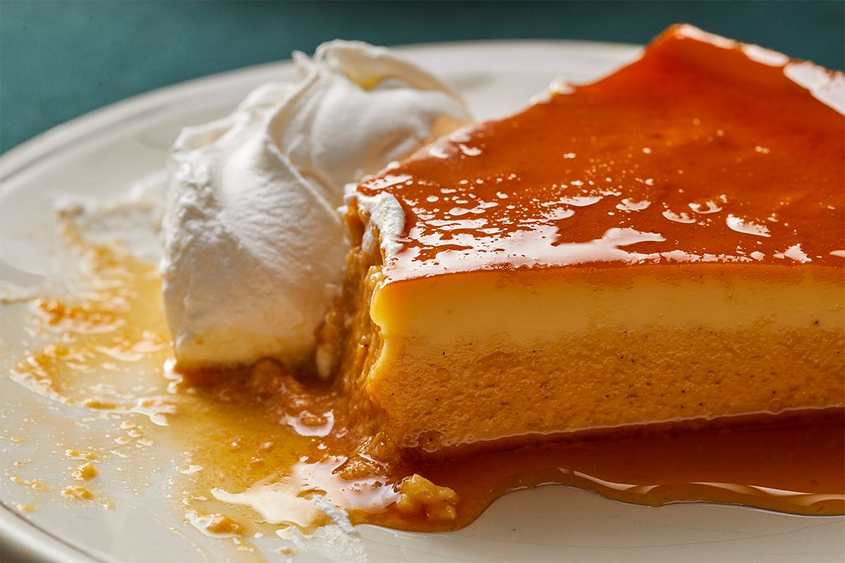 A close-up of a slice of flan topped with golden caramel sauce, served with a dollop of whipped cream on a white plate.