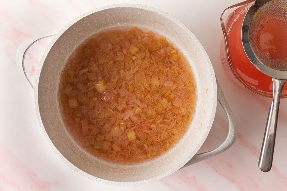 A white pot filled with chopped rhubarb simmering in pink liquid sits on a light surface. Next to it is a glass measuring cup with pink liquid and a metal strainer on top.