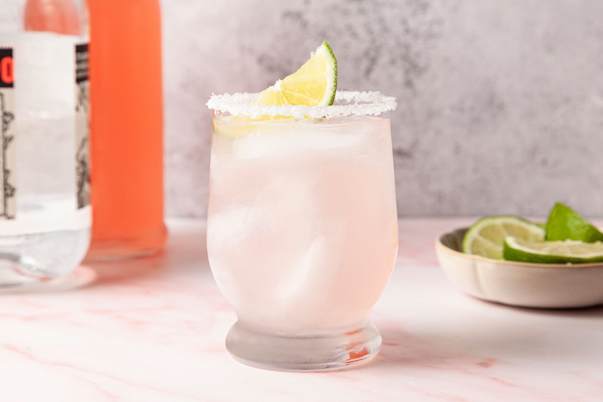A glass of ice-filled pink cocktail with a salted rim and a lime wedge garnish sits on a light marble surface. A bowl of lime slices and bottles are in the background.
