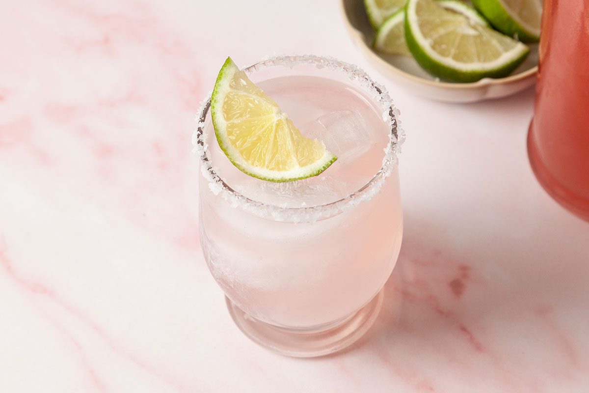 A glass of pink cocktail with a salted rim and a lime wedge garnish, sitting on a light pink marble surface. A bowl of sliced limes is in the background.