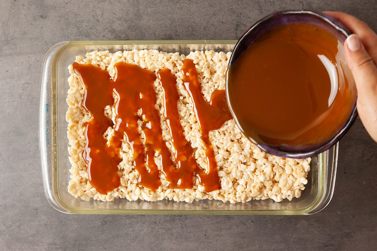 A hand pours caramel sauce from a bowl over a layer of Rice Krispies treats in a glass baking dish on a gray countertop.