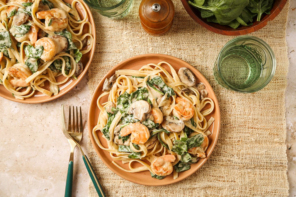 A plate of creamy shrimp pasta with spinach and mushrooms on a woven placemat, accompanied by a fork, knife, glass of water, salad bowl, and pepper grinder. Another plate of pasta is partially visible.