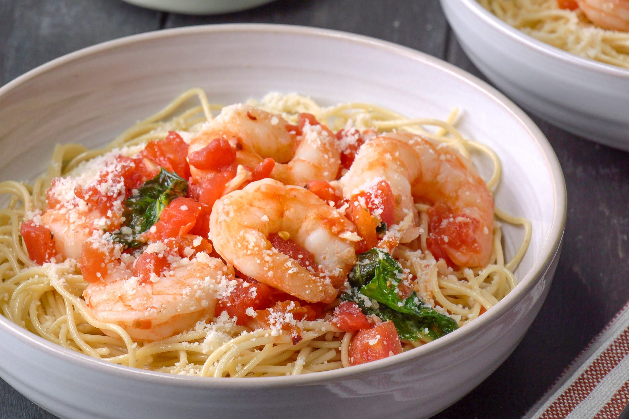3/4 angle view shot of Shrimp Pomodoro served over spaghetti and topped with cheese in two bowls; forks rest on a folded napkin nearby; all set on a black wooden surface