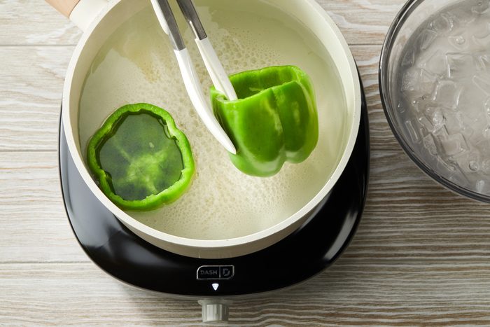 Overhead shot of a skillet over a induction; blanch peppers in boiling water for 5 minutes; Drain and rinse in cold water; set aside; tongs; ice water glass bowl; all set on a cream-colored wooden surface;