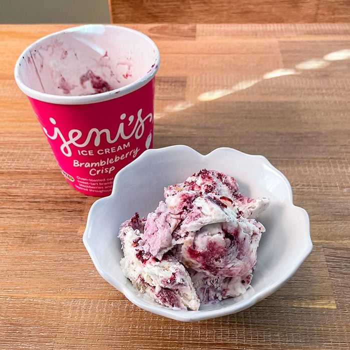 A white bowl filled with scoops of berry swirl ice cream sits on a wooden surface next to a pink Jeni’s Ice Cream container labeled 