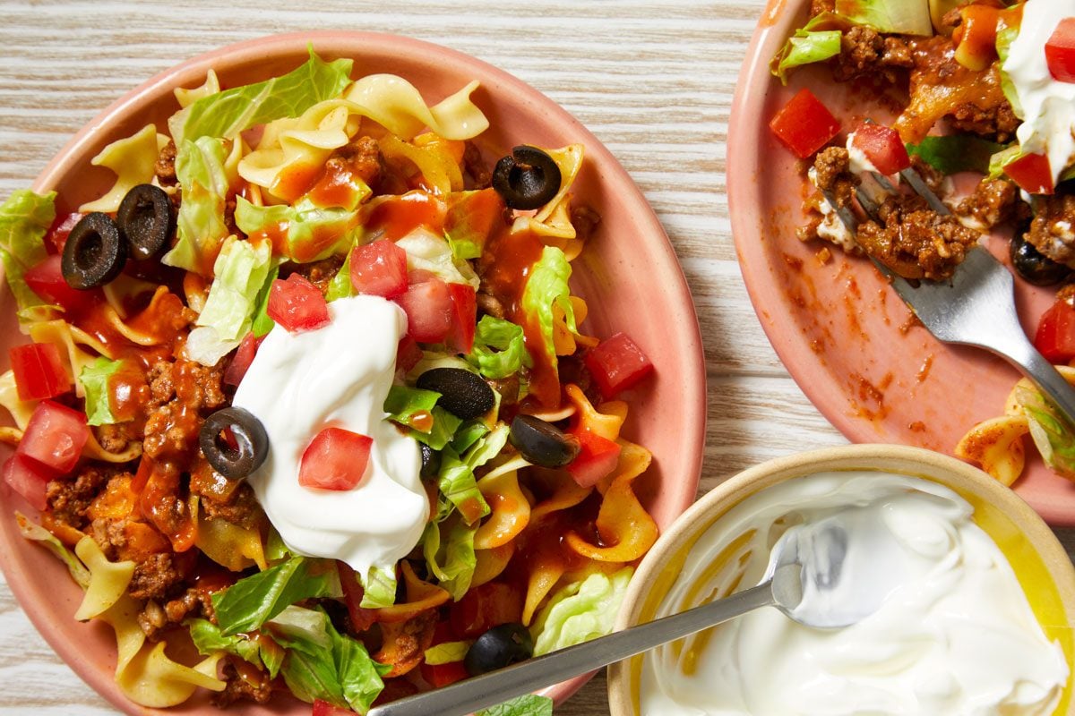 Close-up shot of Taco Noodle Bake in a baking pan; Top with lettuce; tomatoes; olives and taco sauce; a portion served on two small plate garnished with sour cream; a fork; set on a cream-colored wooden surface;