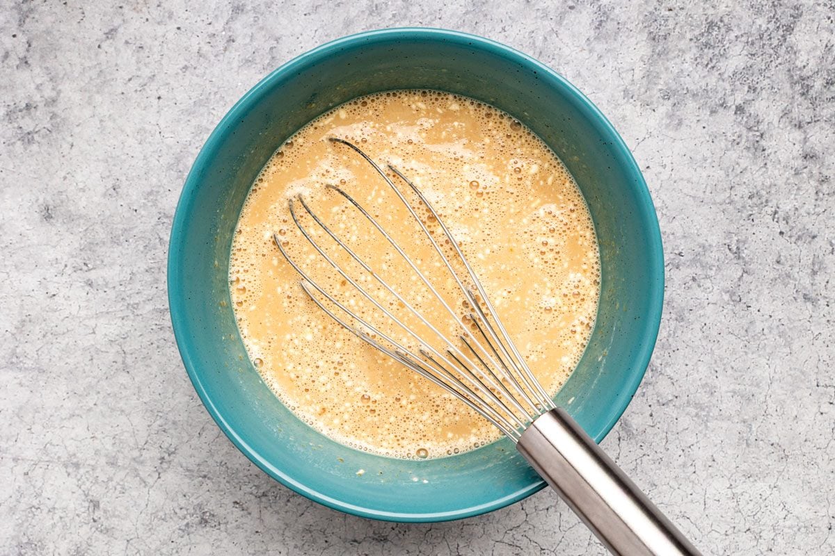 Overhead shot of a bowl combine apple butter; egg; sugar; flour and milk; mix well with whisk tool; on a grey marble surface;