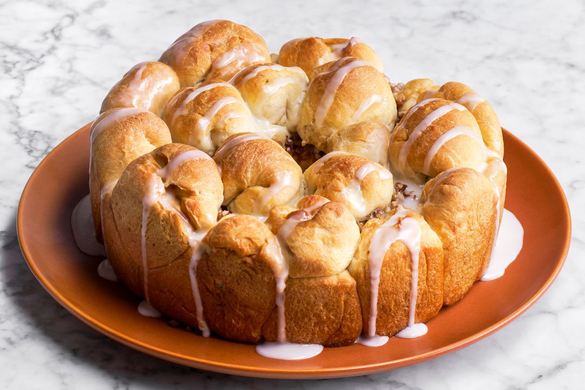 3/4th shot of a round pull apart apple bread ring with golden brown rolls, drizzled with white icing, sits on an orange plate atop a marble surface