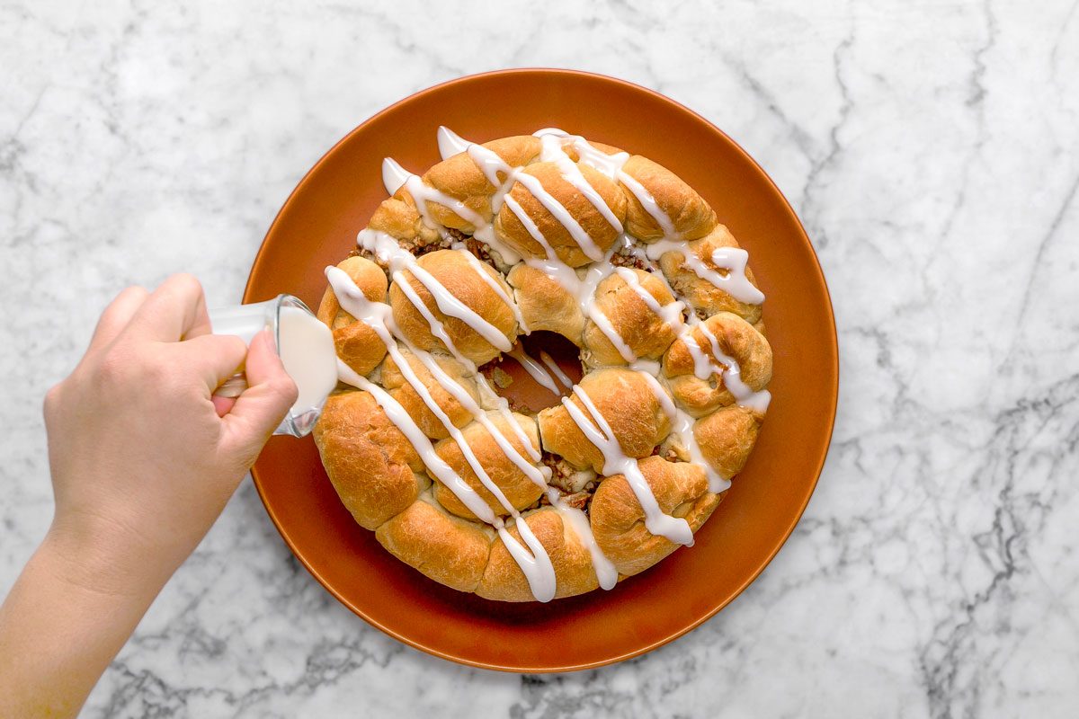 overhead shot of a hand drizzles white icing over a ring shaped pull apart bread on a brown plate, set on a marble surface